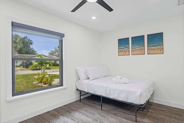 Une chambre avec un lit, une fenêtre donnant sur une maison et de la verdure, et trois œuvres d’art mural sur le thème de la plage.