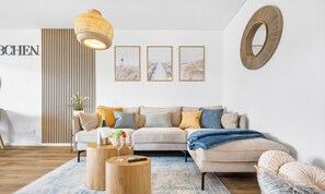 A modern living room with a sofa, coffee table, and a hanging light fixture. - Strandkörbchen - Infrared heat cabin, fireplace, sun loungers, fly screens (Olpenitz)