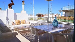 Een buitenterras met een tafel en stoelen, een witte bank met decoratieve kussens en een blauwe decoratieve vaas.