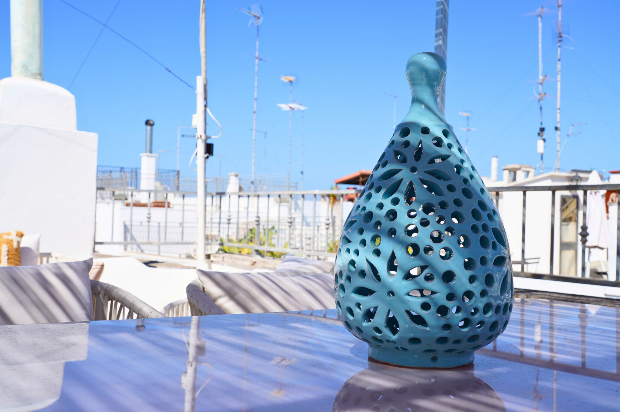 A blue perforated vase on a table with a clear view of the surrounding area, including a railing and some buildings in the background.
