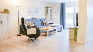 A modern living room with a sofa, a chair, a coffee table, a floor lamp, and a cabinet. - Coastal berth - KÜSTE33/14 - Sauna. Balcony. Family-friendly. (Tossens)