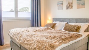 A bedroom with a bed, curtains, a nightstand, and wall art. - Coastal berth - KÜSTE33/14 - Sauna. Balcony. Family-friendly. (Tossens)
