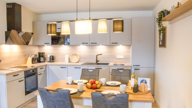 A modern kitchen with white cabinets, a wooden dining table, and a fruit bowl. - Coastal berth - KÜSTE33/14 - Sauna. Balcony. Family-friendly. (Tossens)
