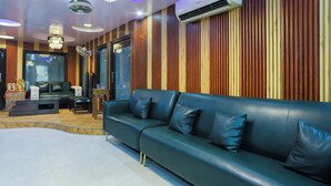 A modern living room with a dark leather sofa, a chandelier, and a large window. - Anand Hotel With banquet hall Karol Bagh (New Delhi)