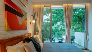 A hotel room with a bed, a view of trees through large windows, and a balcony with chairs. - Palette by RÊVE Escapes - Premium 2 BHK apartment Near Anjuna Beach (Assagao)
