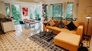 A modern living room with a sofa, coffee table, and dining area. There is a large window with curtains, a ceiling fan, and a clock on the wall. - Palette by RÊVE Escapes - Premium 2 BHK apartment Near Anjuna Beach (Assagao)