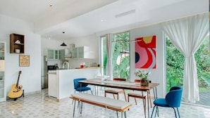 A modern kitchen with white cabinets, a dining area with a wooden table and blue chairs, and a guitar on the floor. - Palette by RÊVE Escapes - Premium 2 BHK apartment Near Anjuna Beach (Assagao)