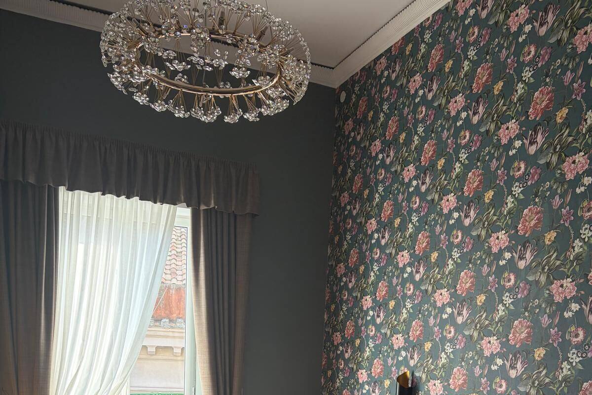 Une chambre avec un grand lit, un papier peint à motifs floraux, un lustre et une vue sur un bâtiment à l’extérieur.