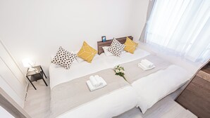 Una cama bien hecha con sábanas blancas, una mesita de noche con una lámpara y una ventana con cortinas de encaje.