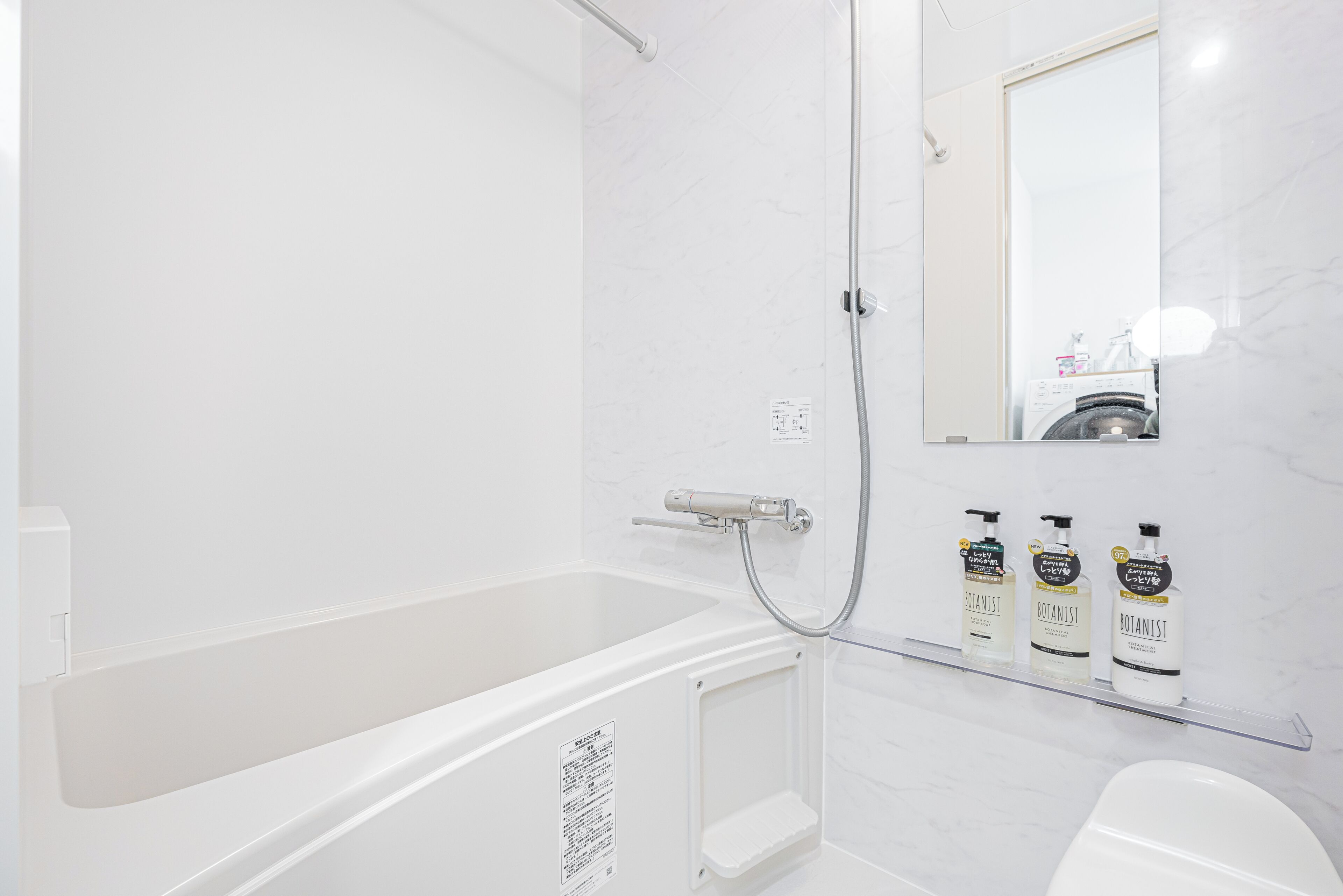 Ein modernes Badezimmer mit einer Badewanne, einer Dusche und einem Spiegel. Auf einem Regal befinden sich drei Flaschen 'Botanist' Shampoo.
