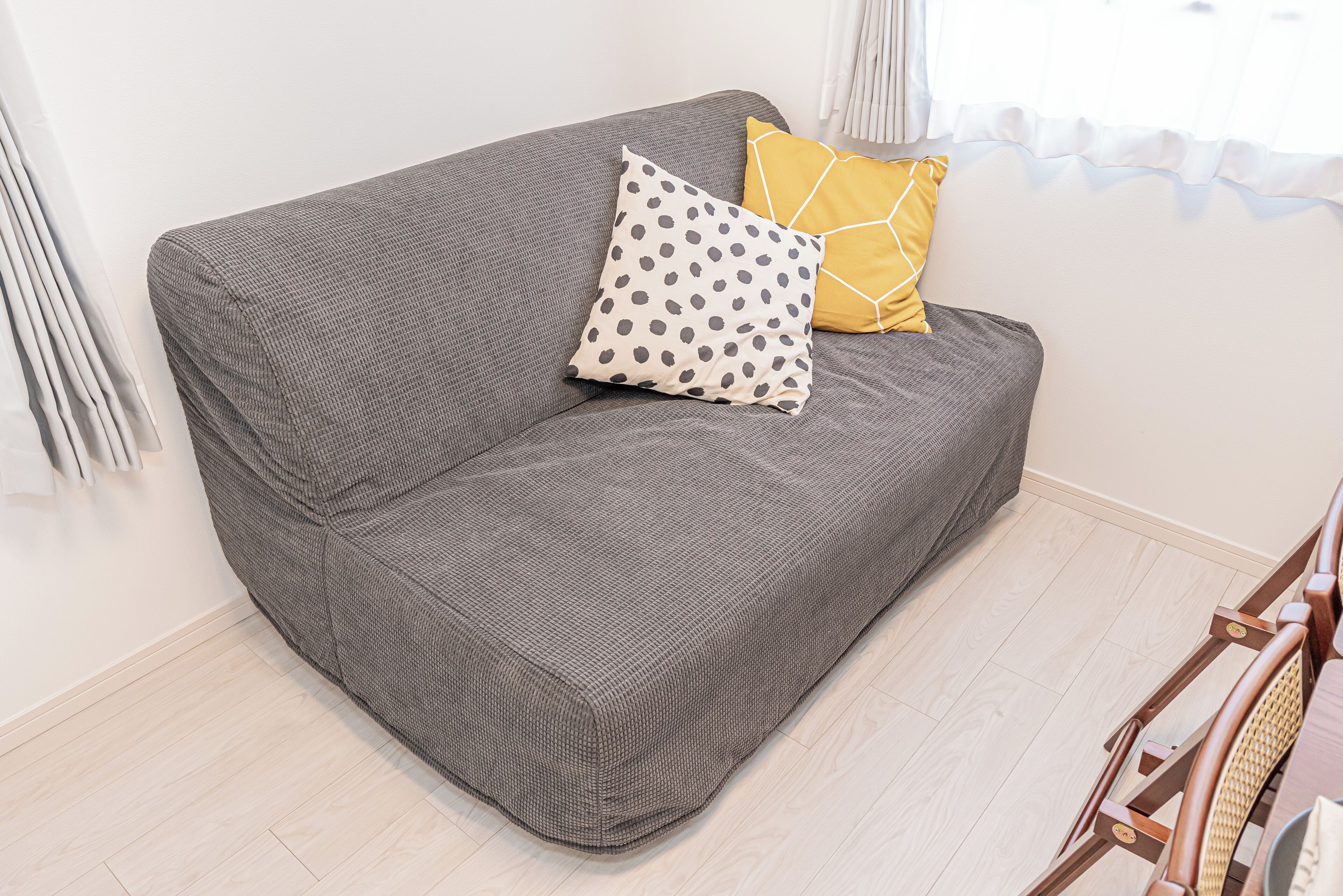Un canapé gris recouvert d’un tissu texturé, deux coussins (l’un à motifs à pois et l’autre à motifs géométriques), et une chaise en bois avec un dossier en osier tressé.