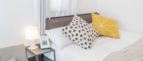 A neatly made bed with a white comforter, two pillows with distinct patterns, a bedside lamp, and a small plant on the nightstand.