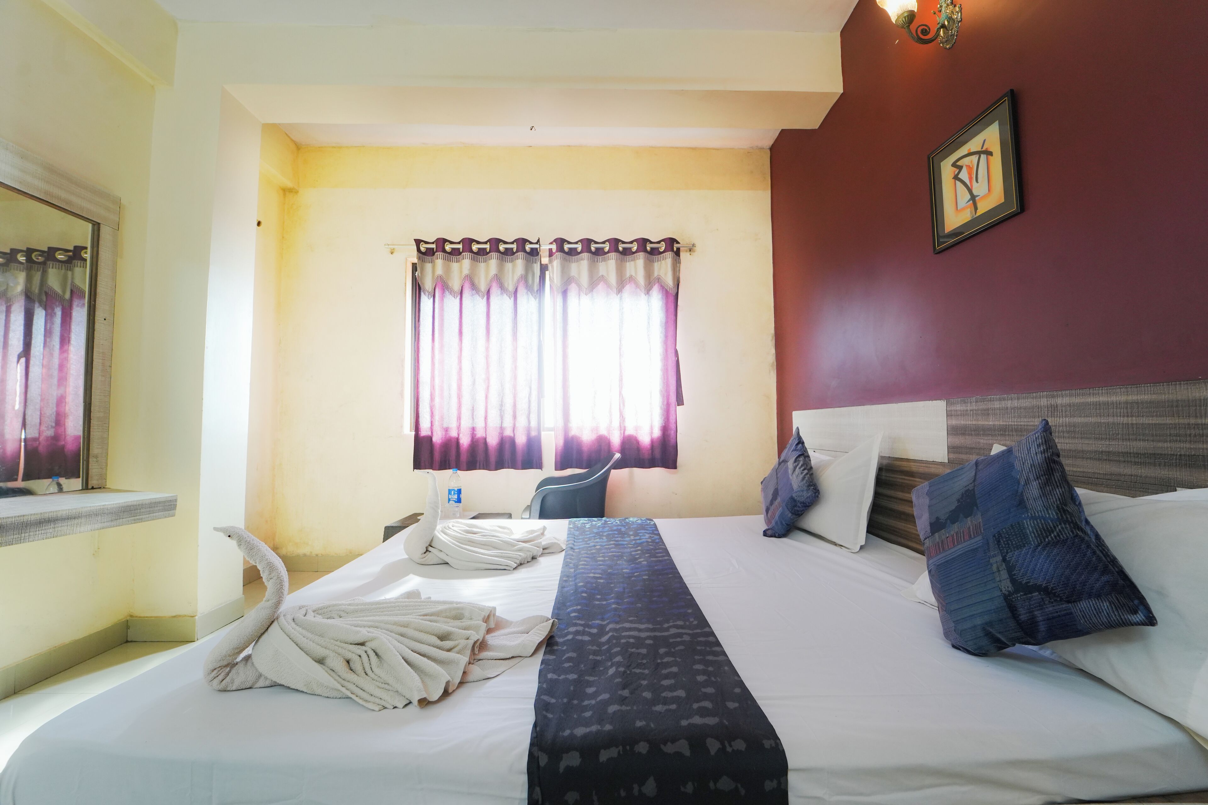 Habitación de hotel con cama, ventana con cortinas, mesita de noche con una botella y un cuadro colgado en la pared.