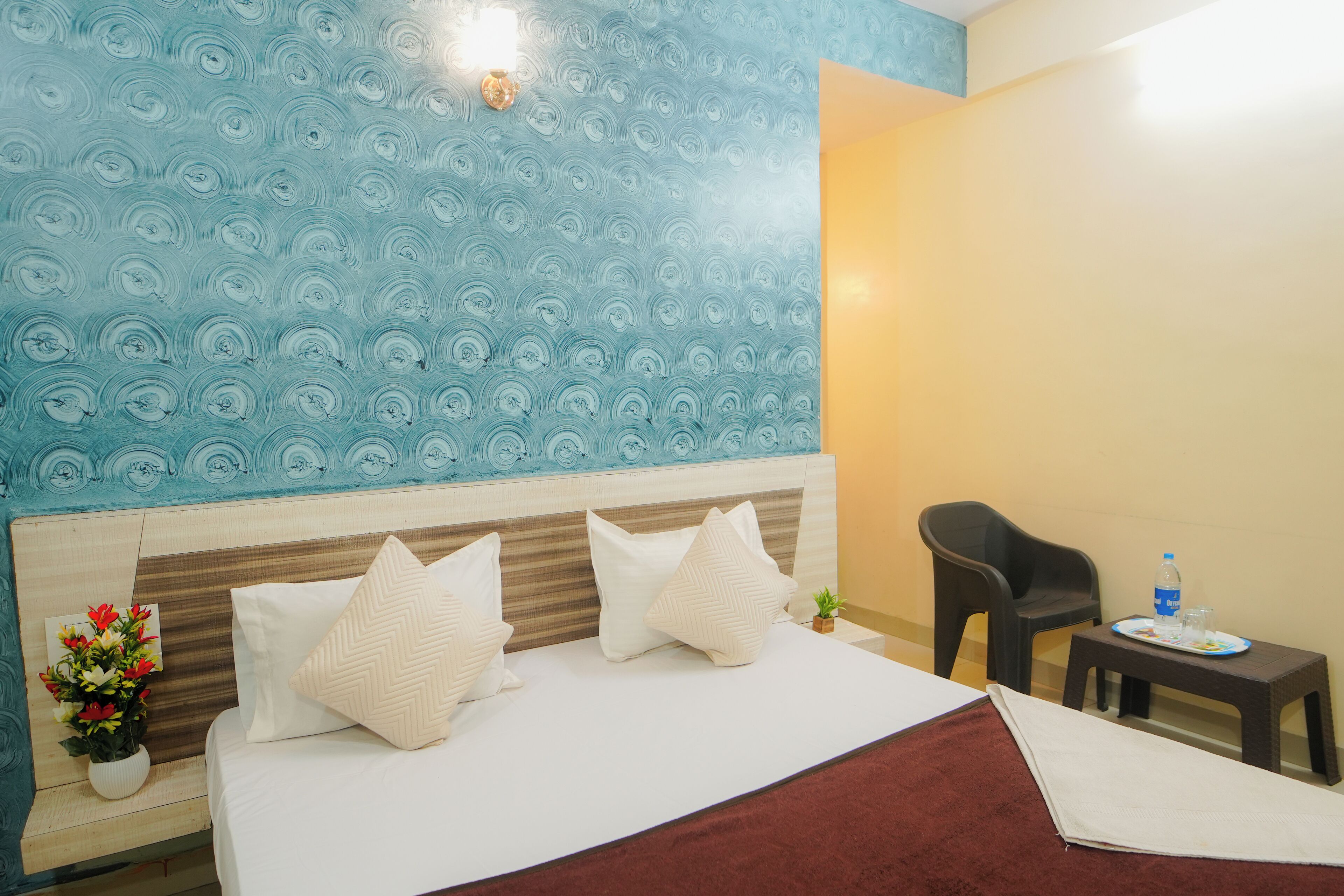A hotel room with a bed, a bedside table with a water bottle and glasses, a chair, and a small potted plant.