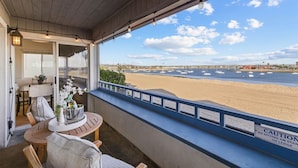 A balcony with a view of the beach and ocean. There is a table with chairs and a potted plant. A caution sign is visible on the railing. - Remodeled 4BR Bayfront Home | Sleeps 12 | Stunning Views + BBQ Patio (San Diego)