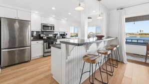 A modern kitchen with white cabinetry, stainless steel appliances, a large island with stools, and a view of the ocean. - Remodeled 4BR Bayfront Home | Sleeps 12 | Stunning Views + BBQ Patio (San Diego)