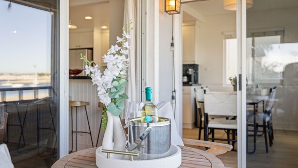A dining area with a round table, a vase with white flowers, and a bottle of wine. - Remodeled 4BR Bayfront Home | Sleeps 12 | Stunning Views + BBQ Patio (San Diego)