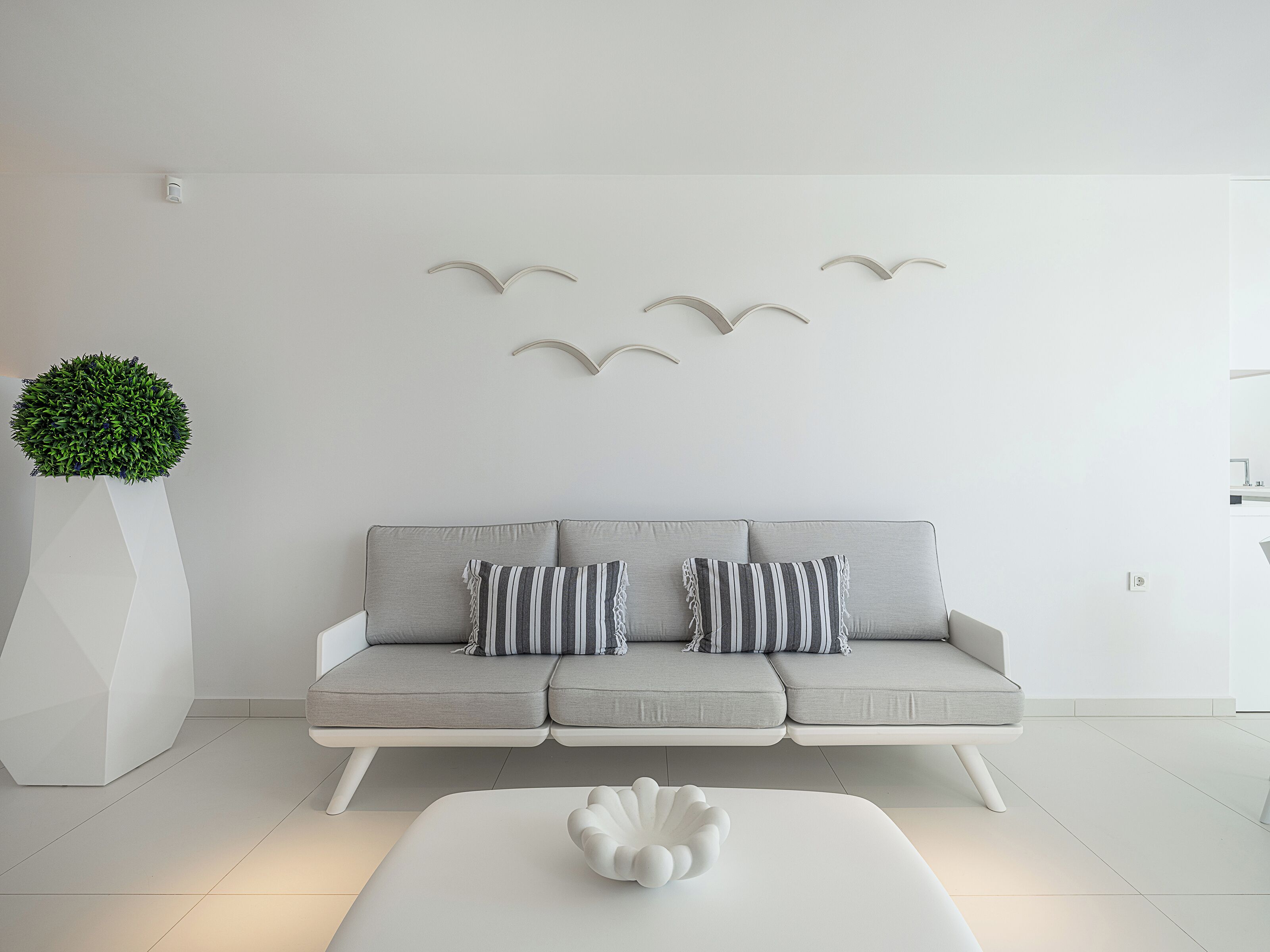 Een moderne woonkamer met een grijze bank, gestreepte kussens, een witte salontafel, een potplant en wanddecoraties van vogels.