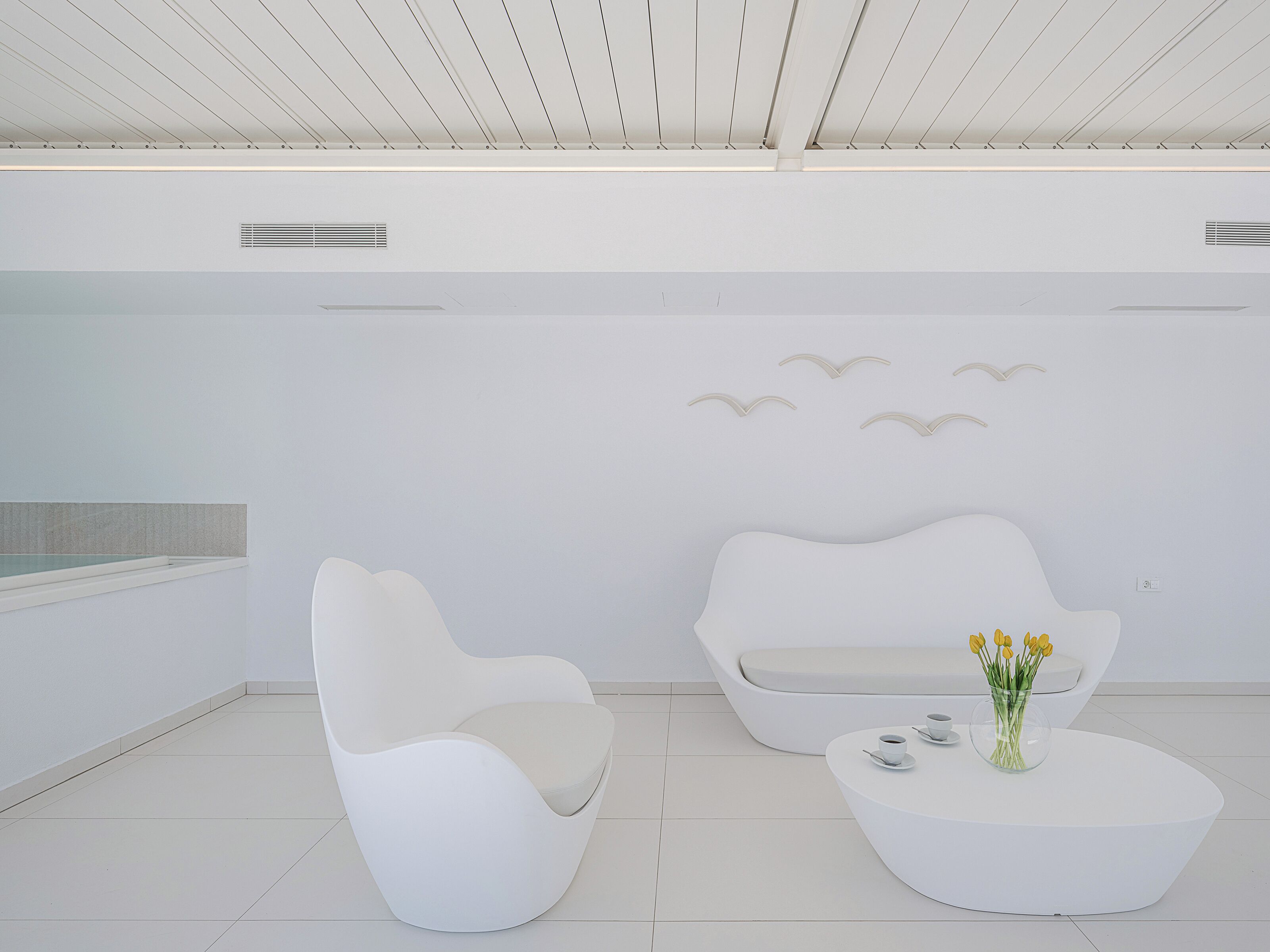 Una habitación moderna y minimalista con una paleta de colores blanca, que presenta un sofá curvo único, una mesa de centro redonda con un florero y una lámpara de pared.