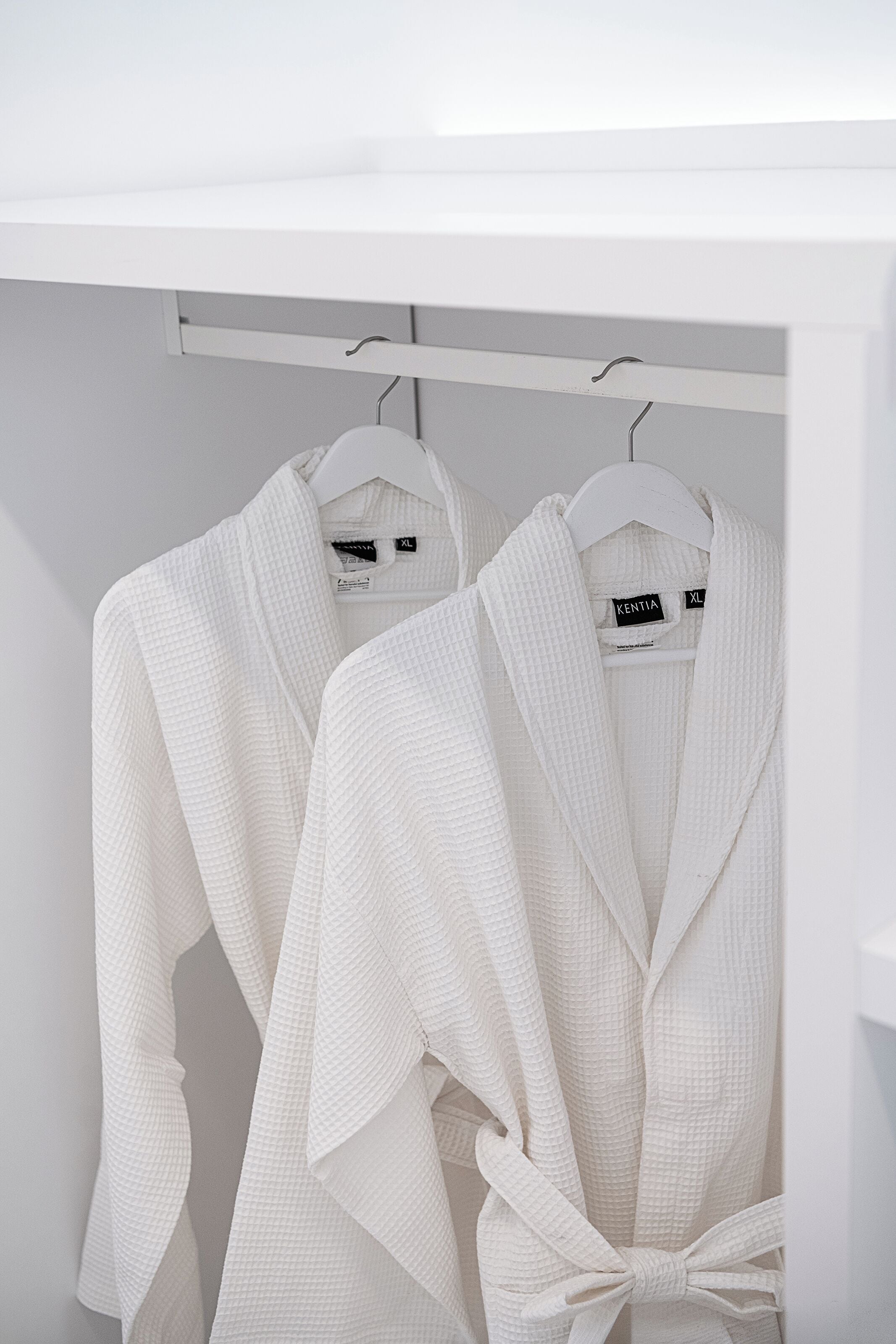 Zwei weiße Bademäntel hängen an einem weißen Kleiderbügel in einem Schrank.