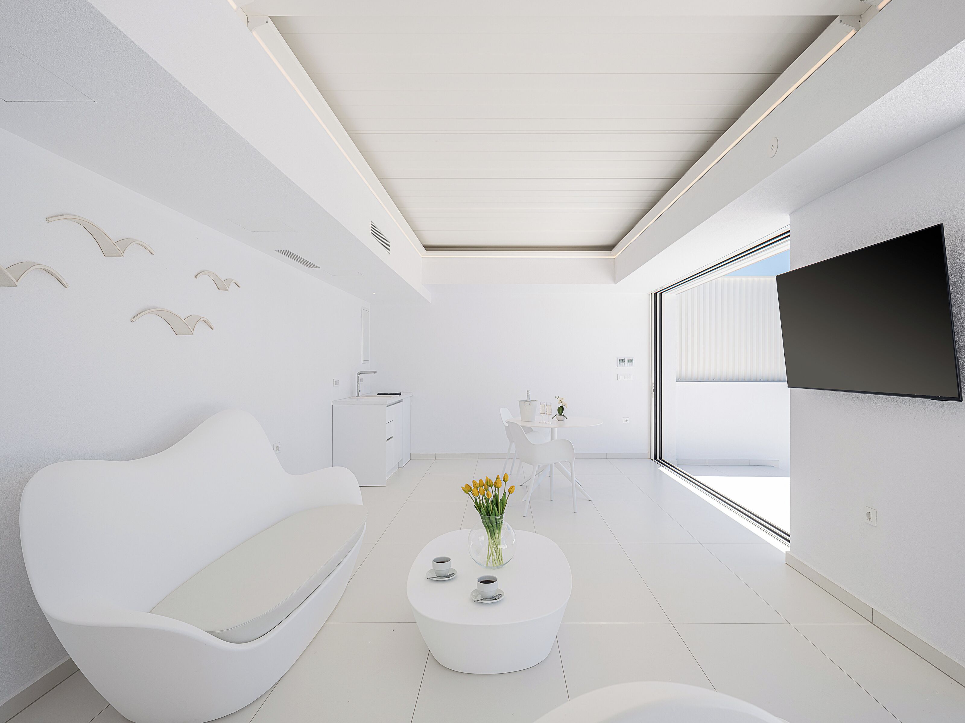 Un salon moderne et minimaliste, avec un canapé blanc, une table basse ornée d’un vase de fleurs, un téléviseur à écran plat fixé au mur et un coin repas comprenant une table et des chaises.