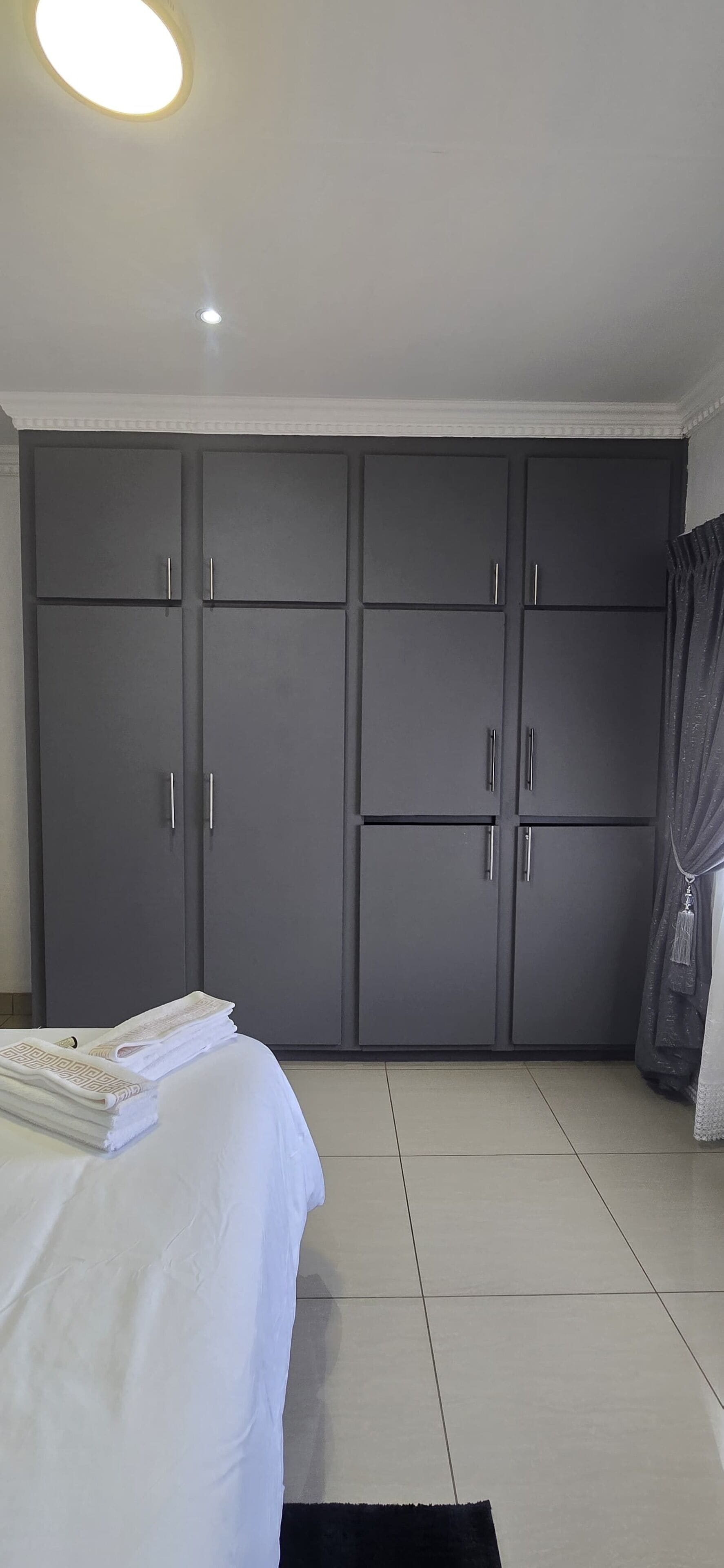 Un dormitorio con un armario grande color gris, una cama con ropa de cama blanca y suelo de baldosas.