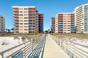 Ein Promenadenweg führt zu Strandapartments mit Balkonen, am strahlend blauen Himmel und feinem Sandstrand.