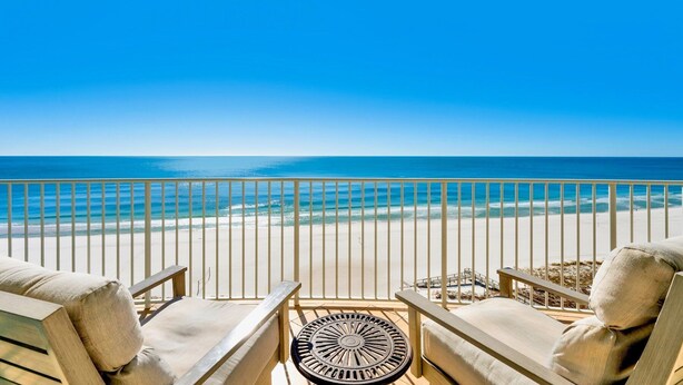 A balcony with two beige cushioned chairs and a round table, overlooking a beach with a clear blue sky and calm sea.