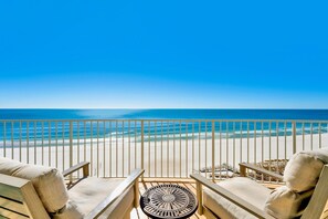 A balcony with two beige cushioned chairs and a round table, overlooking a beach with a clear blue sky and calm sea. - Turquoise Place C1008 | Gorgeous, luxury condo with grill and hot tub on deck! (Orange Beach)