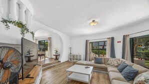 Ein geräumiges Wohnzimmer mit einem weißen Sofa, einem weißen Couchtisch und einem großen Ventilator. Im Hintergrund ist ein Balkon mit Pflanzen und ein Essbereich zu sehen.