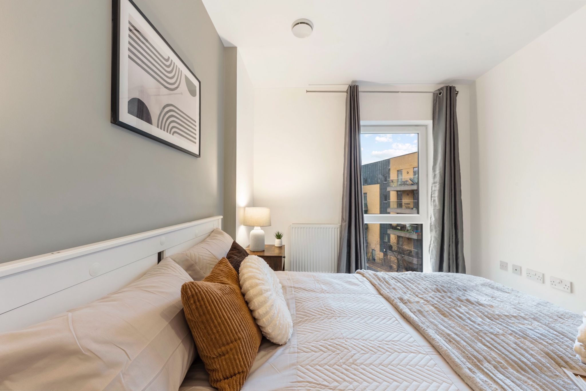 A neatly made bed with a patterned bedspread and pillows, a framed artwork on the wall, a bedside lamp, and a view of the outside through a window with curtains.