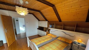 Quarto com teto inclinado, piso de madeira, cama com colcha xadrez, mesa de cabeceira com abajur e prateleiras embutidas.