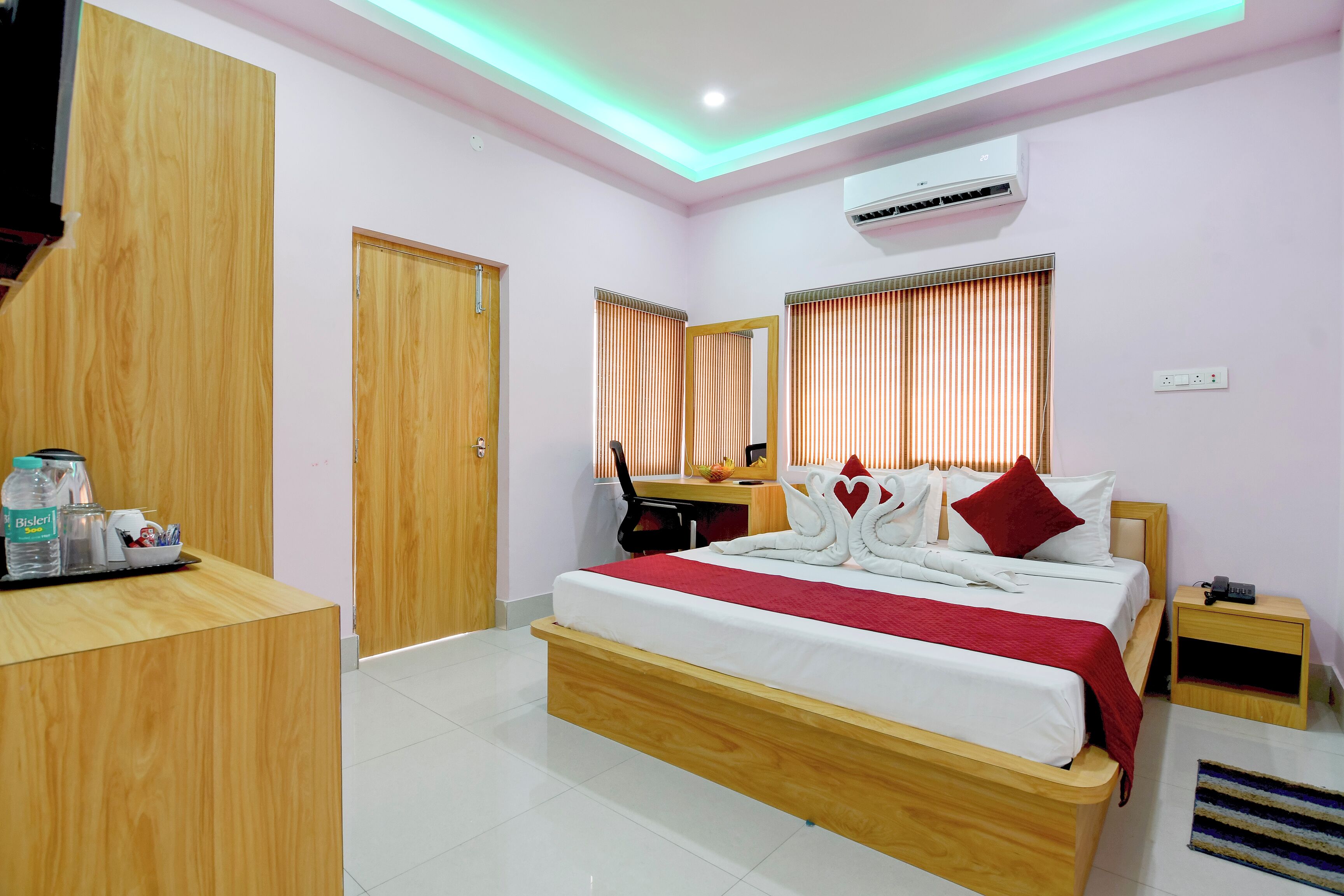 A hotel room with a wooden bed, a desk, a chair, an air conditioner, and a reception area with a counter and some items on it.
