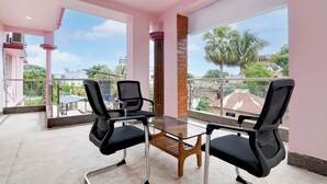 A balcony with two black office chairs and a glass table, overlooking a residential area with palm trees and houses. - Hotel Chaitrangada (Bolpur)