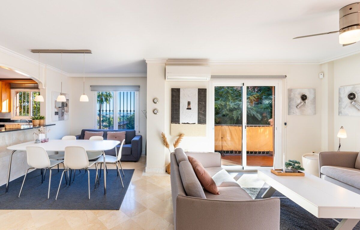 A modern living room with a dining area, a sofa, and a coffee table. There is a sliding glass door leading to a balcony.