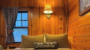 Una habitación acogedora con paredes de madera, una cama con colcha acolchada, un almohadón con la palabra 'Adirondacks', una ventana con cortinas estampadas y una lámpara fijada a la pared.
