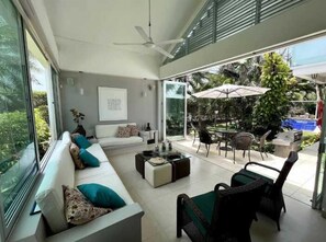 A modern living room with a sofa, coffee table, and chairs. There is a ceiling fan, a dining area with outdoor seating, and a view of greenery outside. - NOK Luxury 4BR Pool Villa in Manzanillo Cartagena (Cartagena)