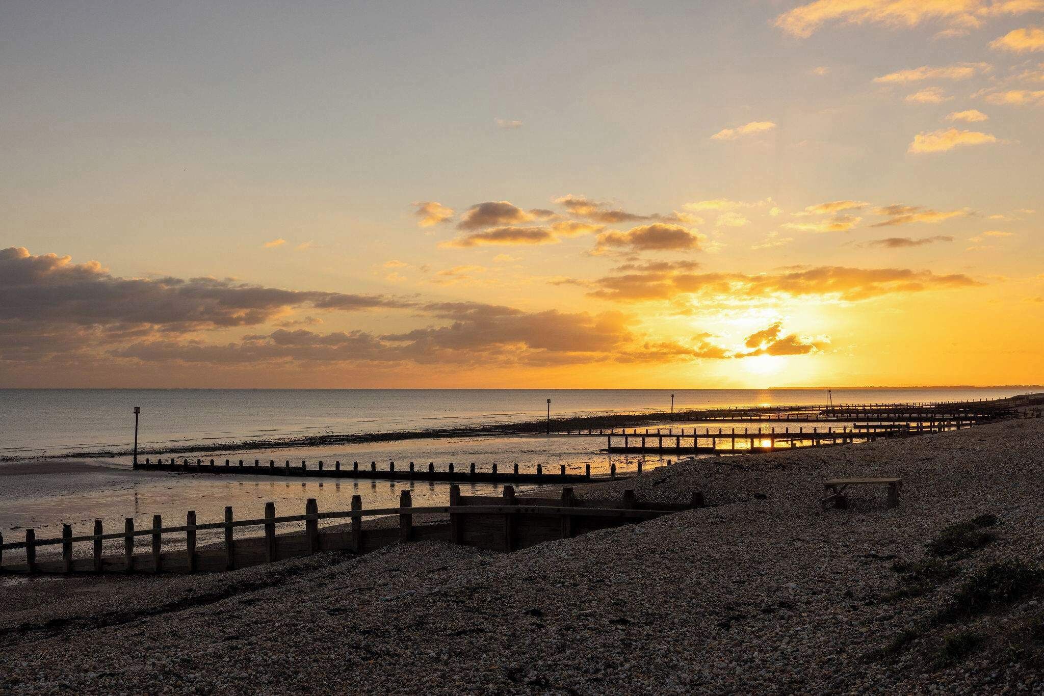 A beach with a wooden pier extending into the water, a bench, and a rocky shoreline.