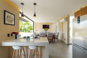 Una cocina moderna con una isla central, taburetes de bar, un comedor con sillas, un refrigerador grande y un televisor montado en la pared.