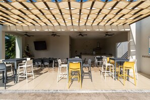 Un patio exterior con mesas y sillas, ventiladores de techo y un televisor montado en la pared.
