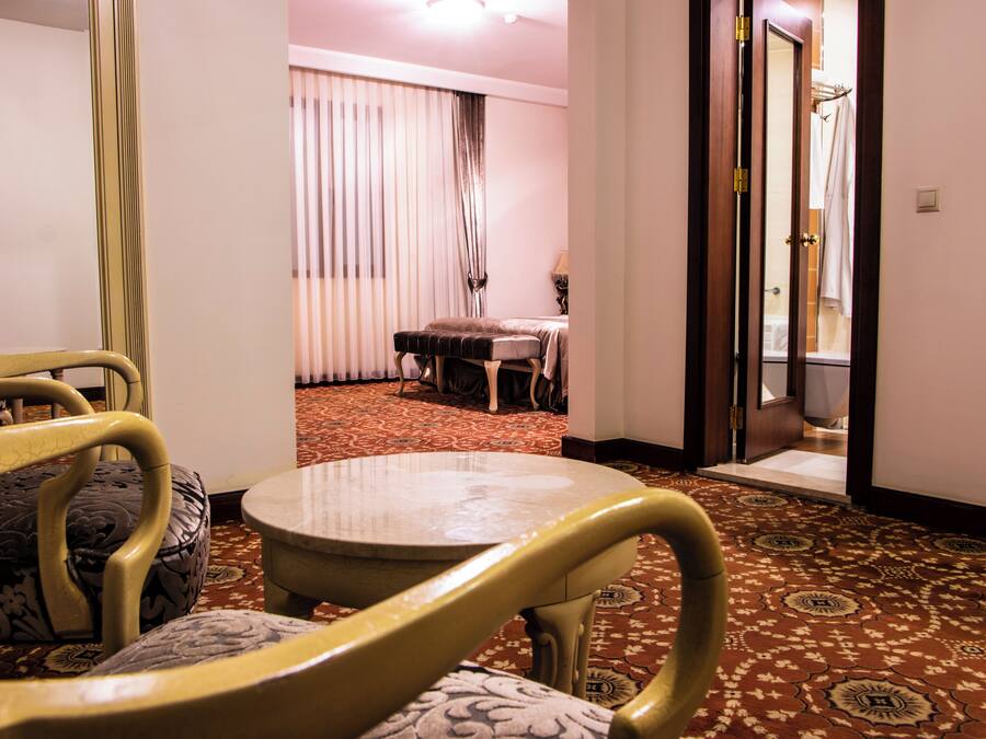 A hotel room with a round table, two chairs, a bench, a bed, and a bathroom visible through an open door.