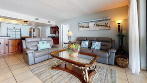 A living room with a glass coffee table, two grey sofas, a wooden coffee table, and a kitchen in the background. - Walkable, All-suite 3br: Suntide III - Unit 1003 (South Padre Island)