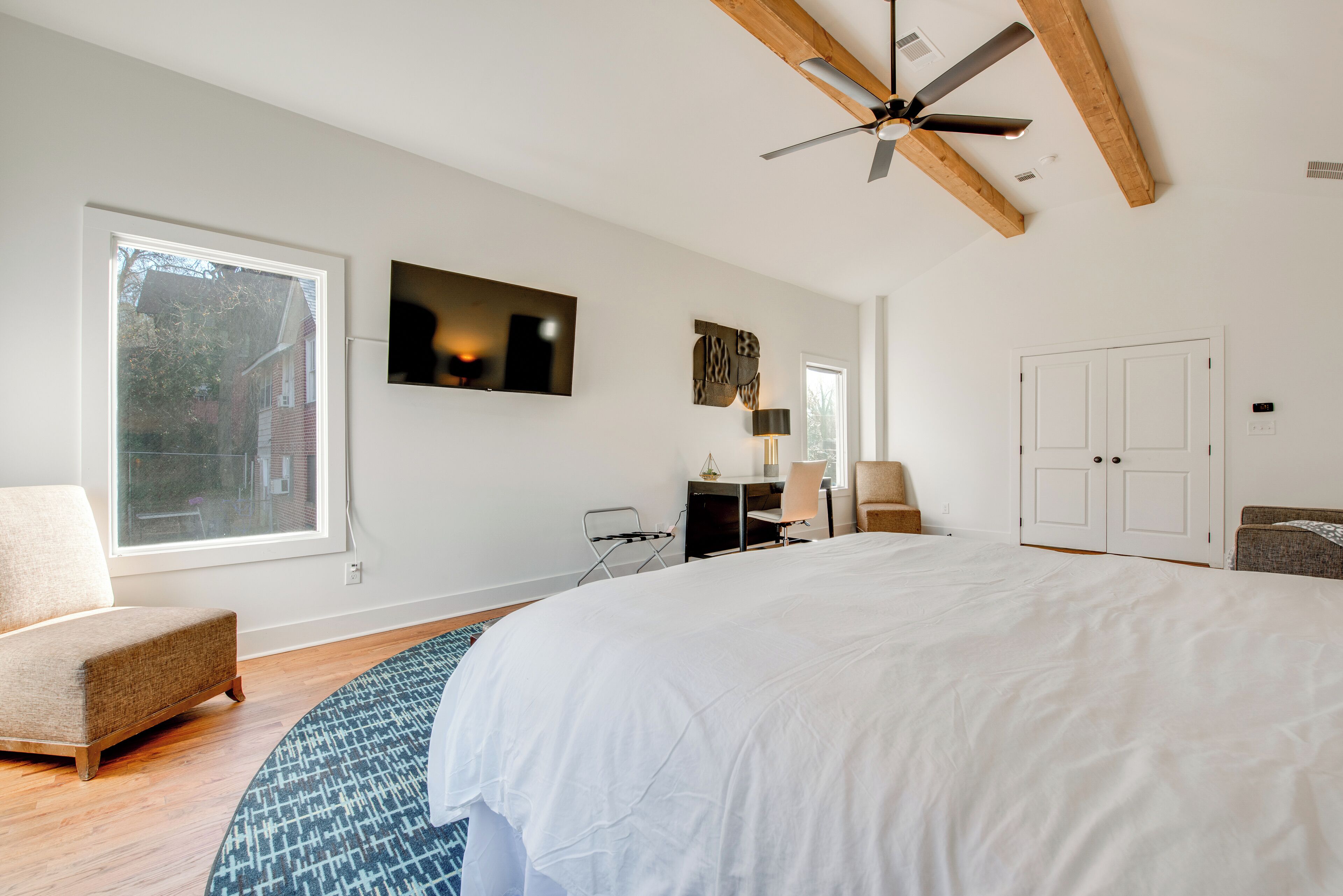 A bedroom with a large bed, a desk, a chair, a ceiling fan, and a view of the outside.