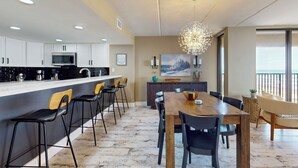 A modern kitchen with white cabinets, a central island with bar stools, a dining area with a wooden table and chairs, and a large window with a view. - Updated 3BR w/ Gulf Views: Suntide III - Unit 801 (South Padre Island)