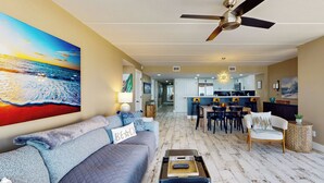 A living room with a sofa, a coffee table, and a painting of a beach scene. There is a ceiling fan and a kitchen in the background. - Updated 3BR w/ Gulf Views: Suntide III - Unit 801 (South Padre Island)