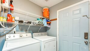 Une buanderie équipée de deux machines à laver et deux sèche-linge, d’étagères contenant des produits de nettoyage et d’une porte bleue.