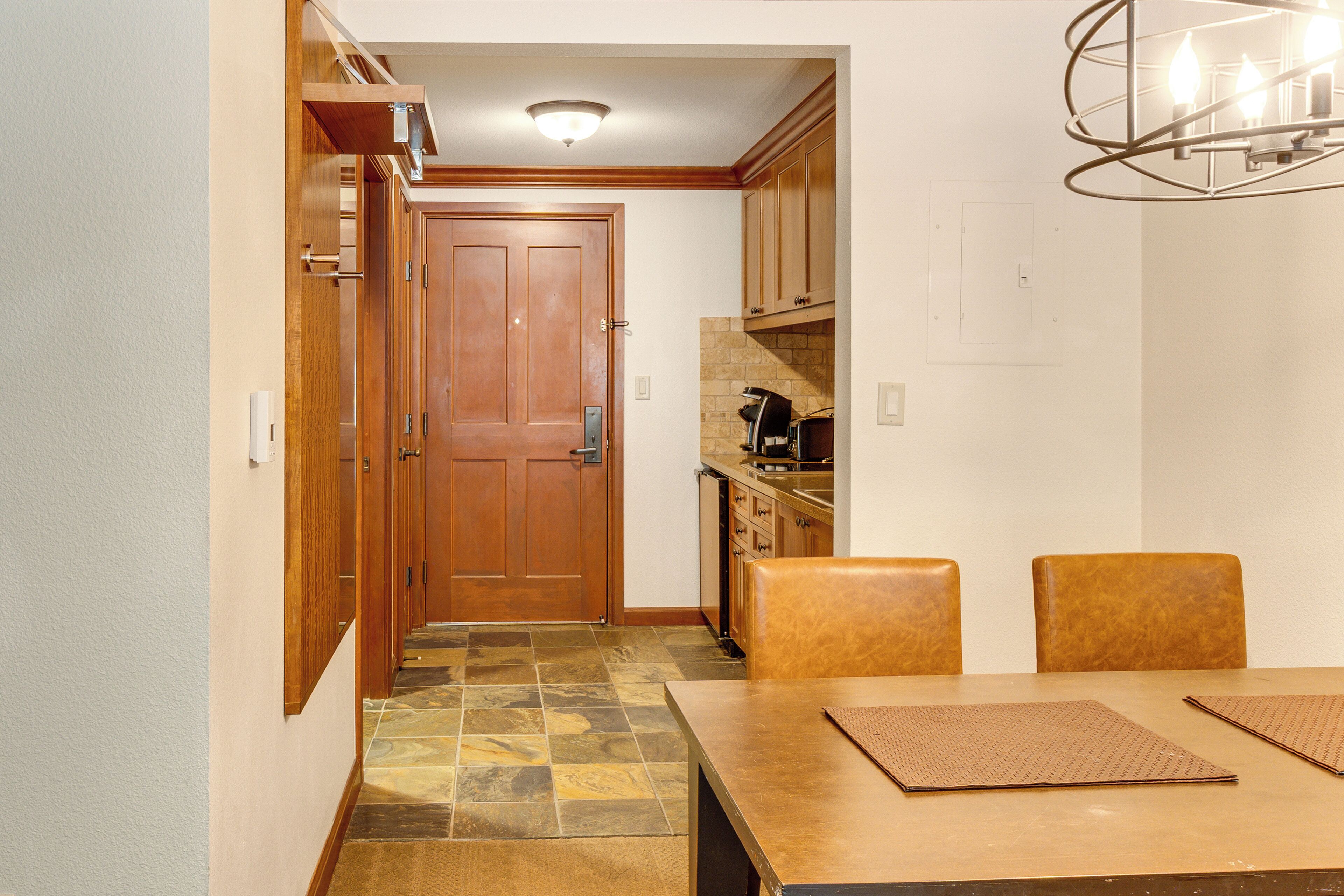 Un pasillo que conduce a una puerta de madera, una zona de cocina con armarios y una mesa de comedor con sillas.