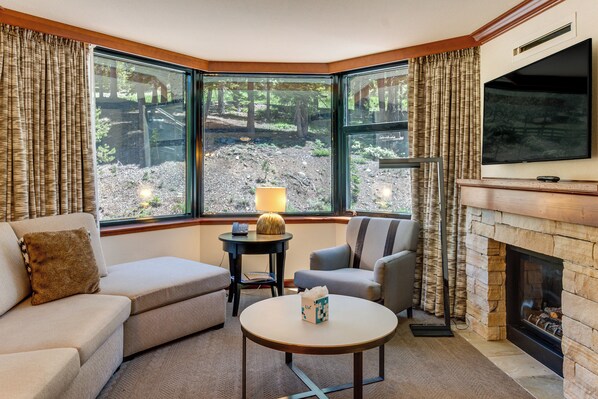 A corner room with a sofa, armchair, and a small table with a lamp. There is a fireplace and a TV mounted on the wall. - 1BR Suite w/ Fireplace, Resort Pools, Spa & Gym (Olympic Valley)