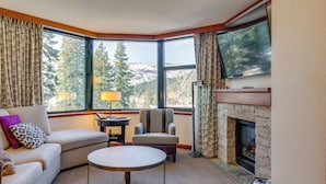 A corner living room with a sofa, armchair, and round table. A fireplace is visible through a window, and a television is mounted above it. - Luxe North Lake Tahoe Getaway in Everline Resort! (Olympic Valley)