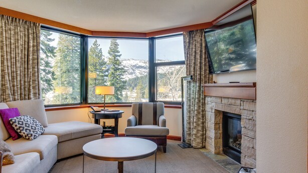 A corner living room with a sofa, armchair, and round table. A fireplace is visible through a window, and a television is mounted above it. - Luxe North Lake Tahoe Getaway in Everline Resort! (Olympic Valley)